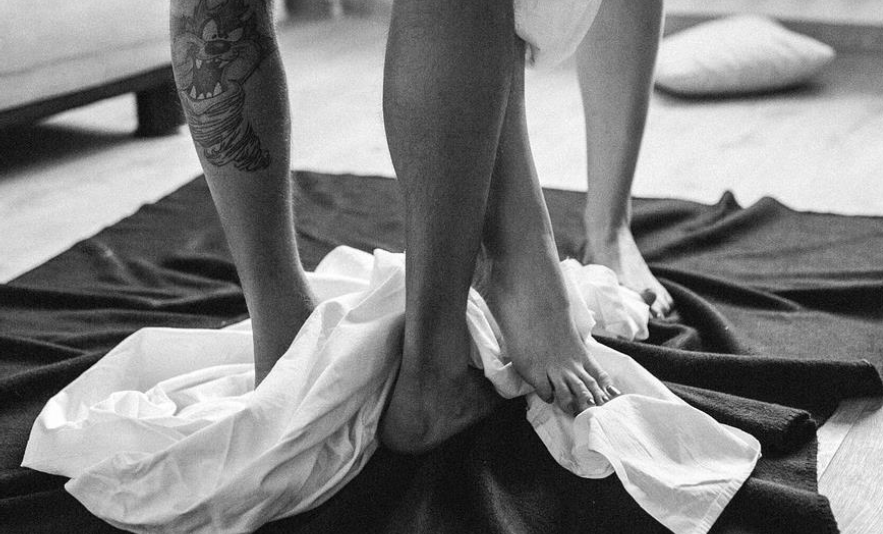 Två par bara ben. De står på en filt och det ligger sägnkläder på golvet. Hon stryker sin vrist mot hans ben. 
