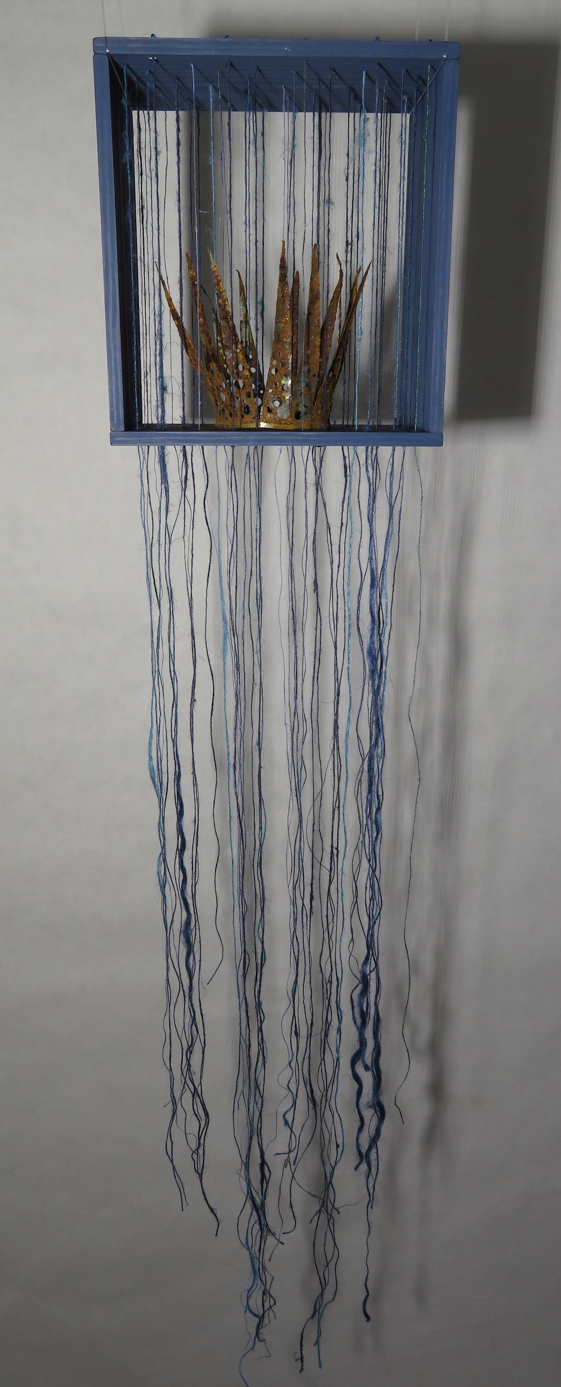 Fotografi av skulptur med en hängande, blå träkub med en krona i metall samt hängande, fria tygtrådar.