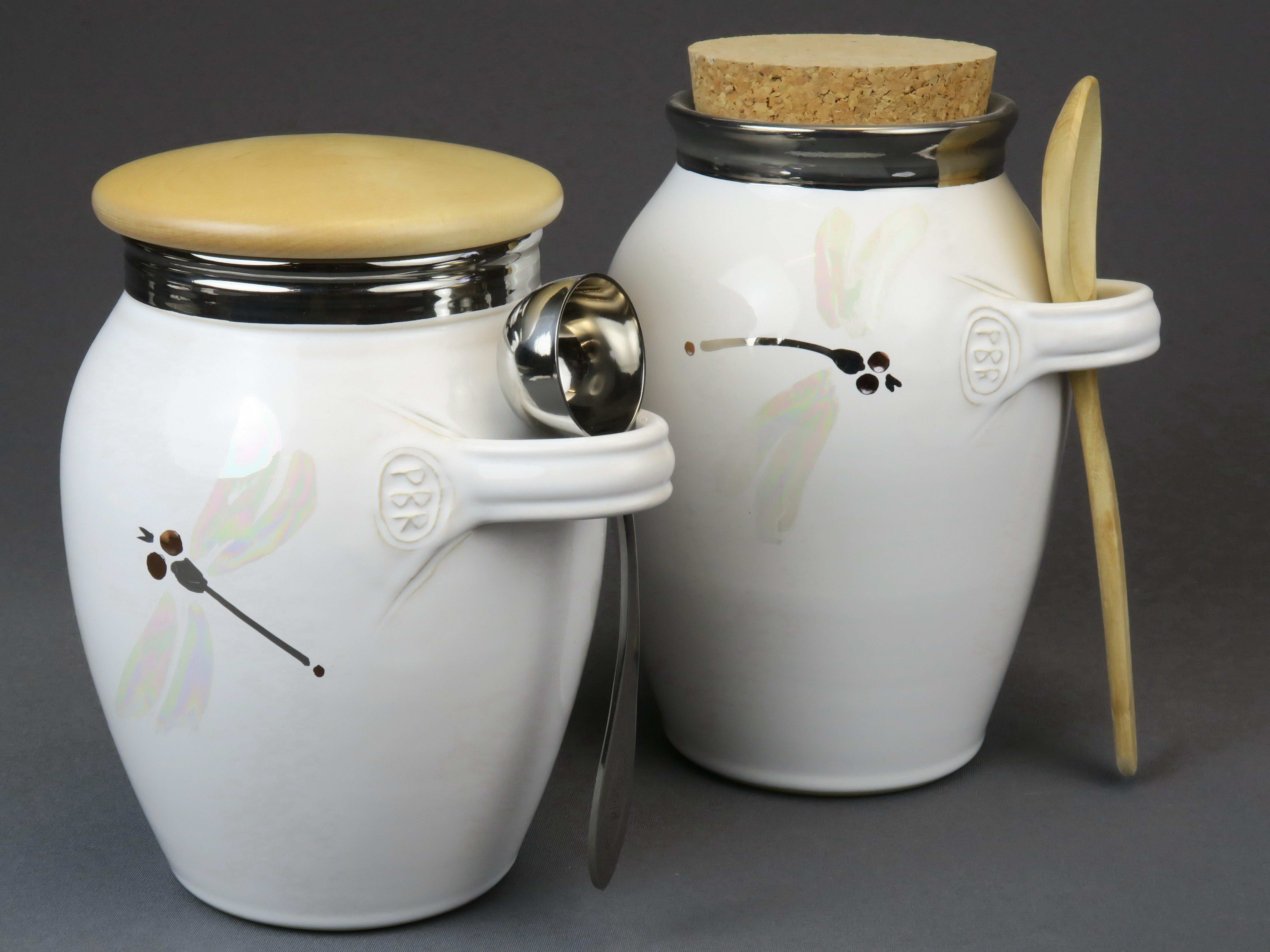 Fotografi av två vita keramikkärl med lock. Små öglor håller varsin sked.