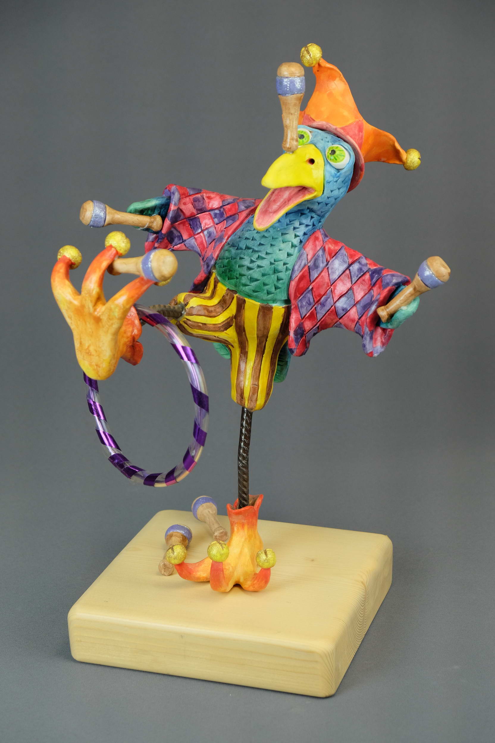 Fotografi av lekfull skulptur av fågel med stora skor, rutig jacka, randiga byxor och bjällermössa. Gul, turkos, rosa, orange.