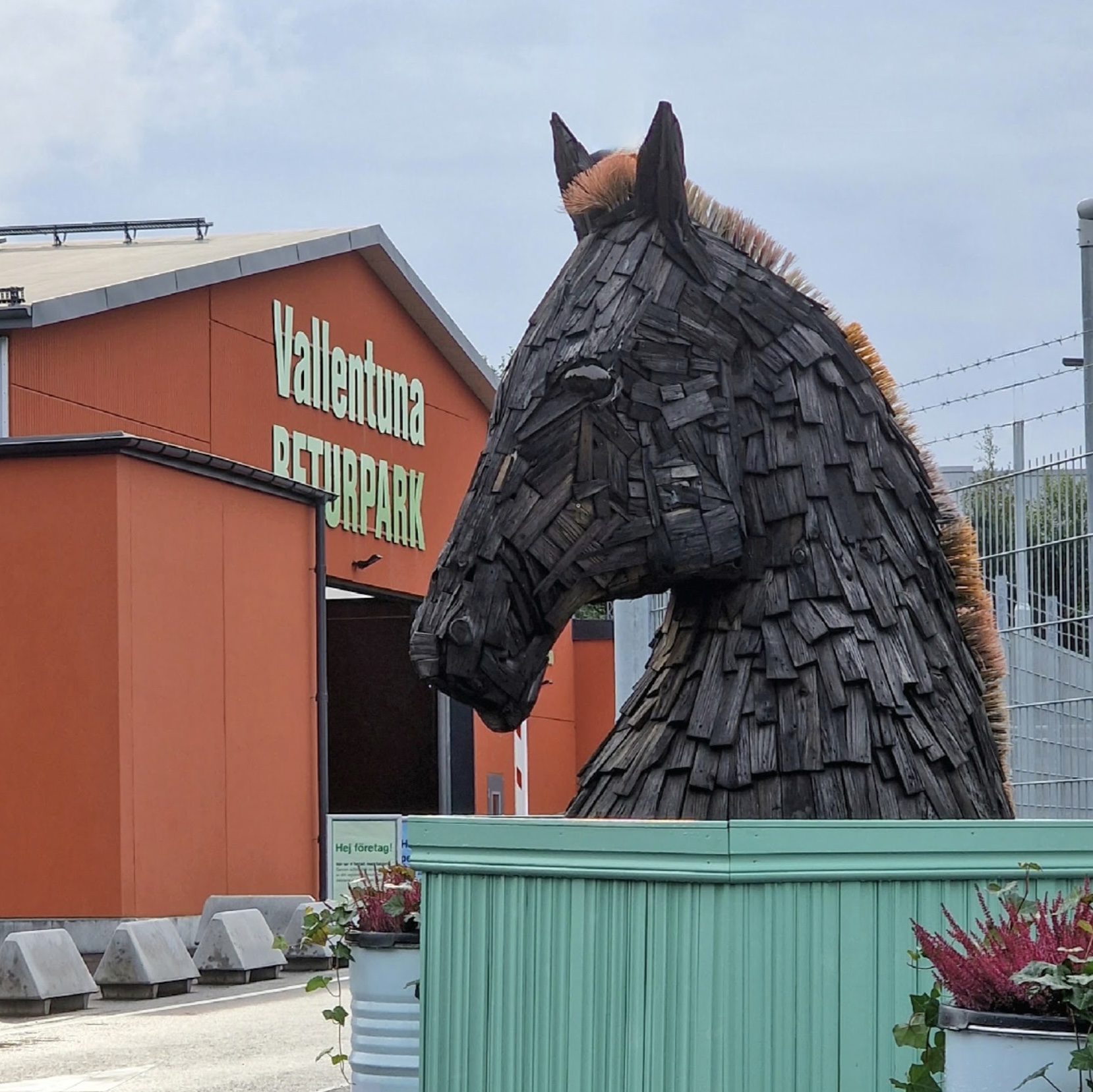 Fotografi av stor skulptur som står utomhus. Skulpturen föreställer ett hästhuvud. Materialet är svarta träbitar och manen är gjord av återbrukade borstar.