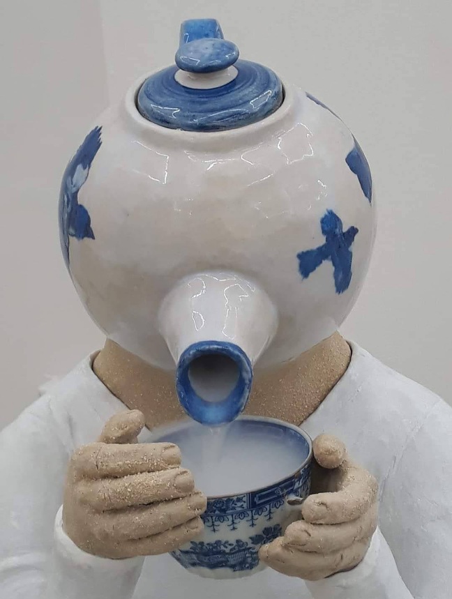Fotografi av keramikskulptur i form av en människokropp med en tekanna som huvud. Vit, hudfärg, blått.