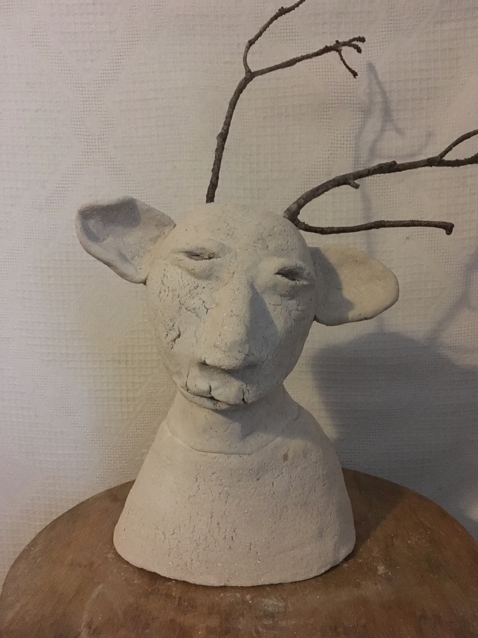 Fotografi av ofärgad, grå lerskulptur. Figur i halvkropp med stora öron och horn av grenar.
