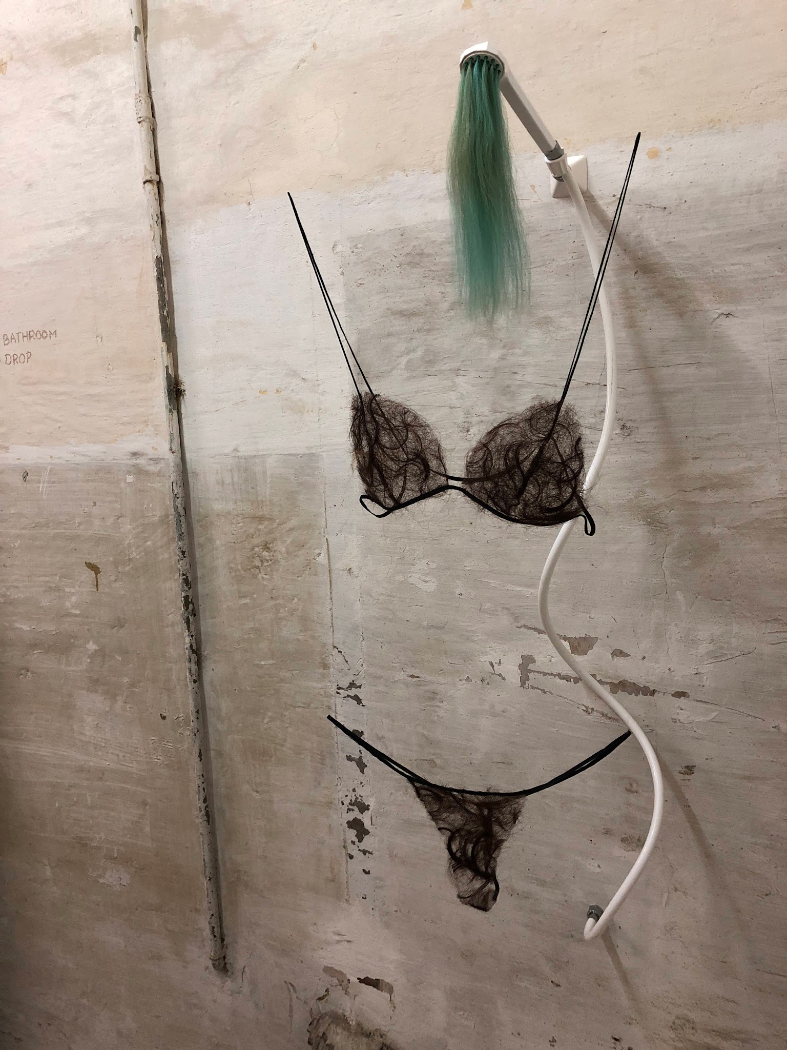 Fotografi av installation med duschmunstycke ur vilket det kommer turkos hår. Under detta hänger en bikini av filtat människohår.