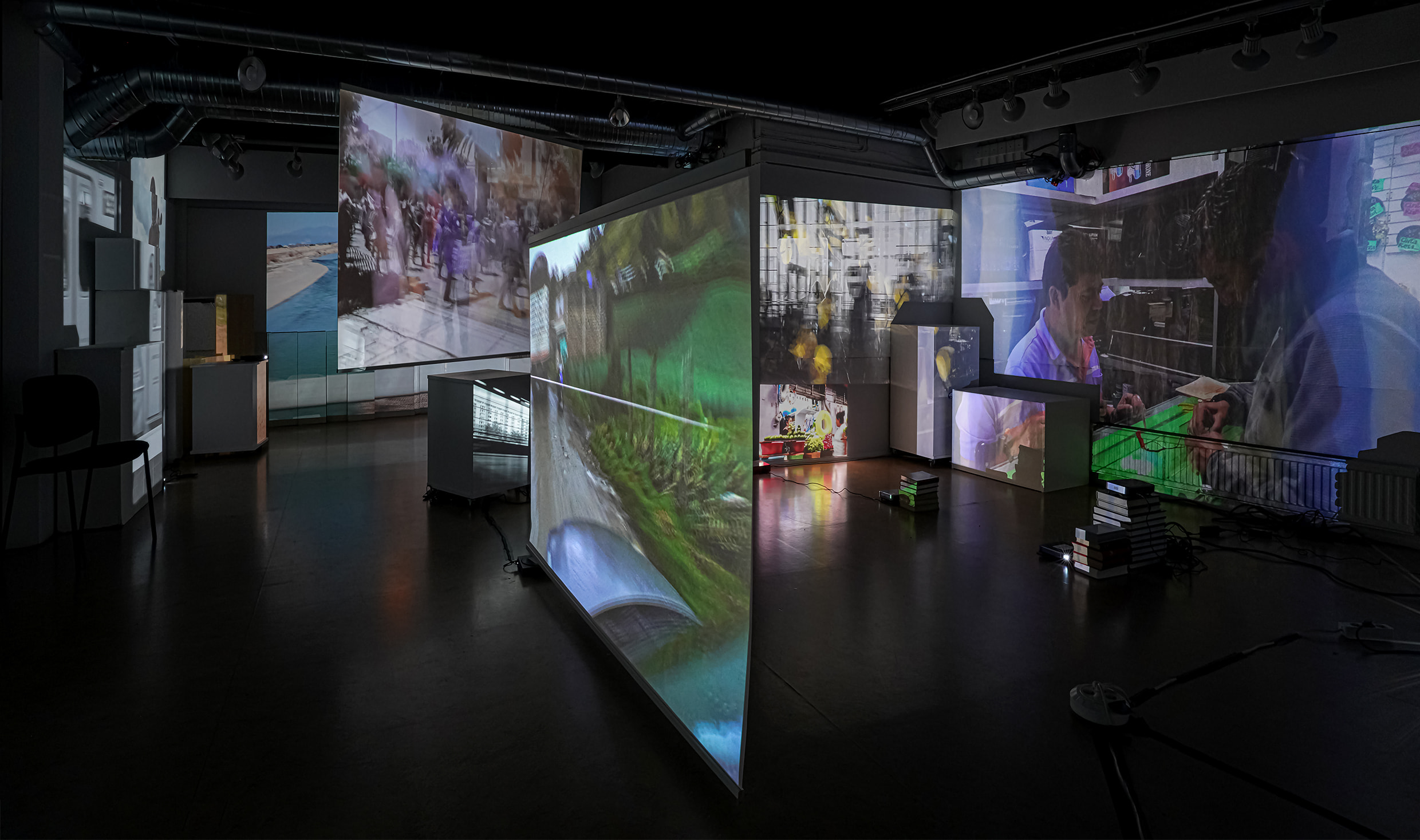 Fotografi av rumslig miljö med flera skärmar på vägg, golv och hängande från tak. Film med människor projiceras på skärmarna.