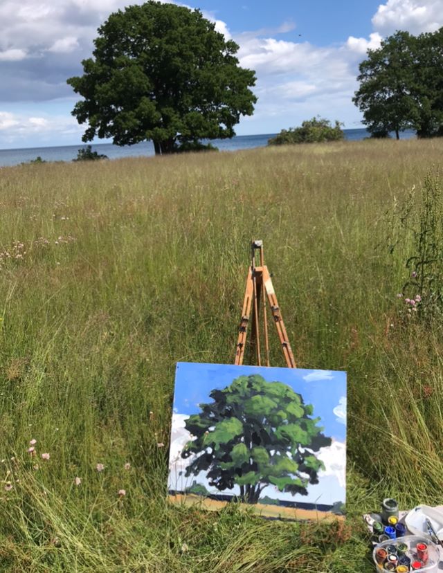 Fotografi av sommarlandskap med träd. På marken står ett staffli och en tavla med en realistisk målning av ett träd i landskap.